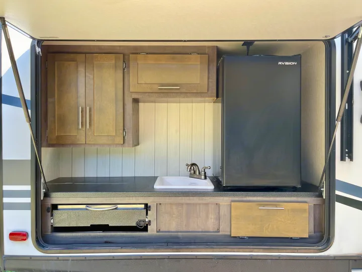 RV rental outdoor kitchen setup with a sink, faucet, mini-fridge, upper cabinets, and a pull-out grill.. Crossroads RV Sunset Trail Super Lite 2019