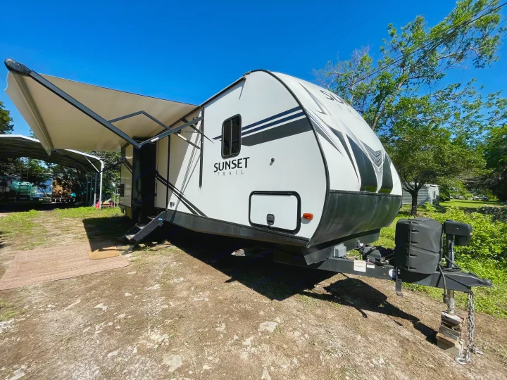This RV rental, a white Sunset Trail travel trailer, is parked with its awning extended under a bright blue sky.. Crossroads RV Sunset Trail Super Lite 2019