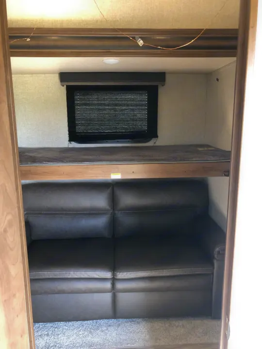 RV rental bunkhouse: A dark grey sofa is positioned under a top bunk, next to a window with a blind. Compact living space.. Keystone RV Cougar 2020