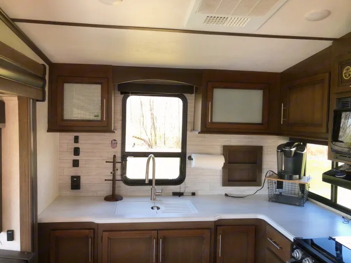 RV rental kitchen with white countertops, modern sink, dark wood cabinets, a coffee maker, and a window overlooking trees.. Keystone RV Cougar 2020