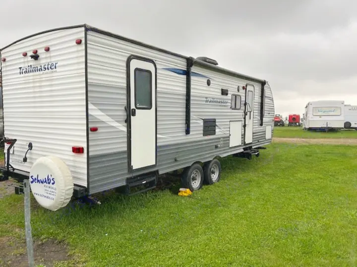 Large, grey and white Trailmaster travel trailer, ideal for an rv rental, on a grassy field. Schwab's RV World tire cover on back.. Gulf Stream Trailmaster 2018