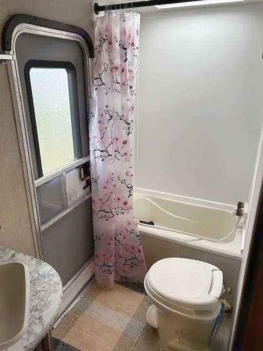 RV rental bathroom interior with pink floral shower curtain, compact tub, and toilet. Features an RV door and vanity.. Gulf Stream Trailmaster 2018