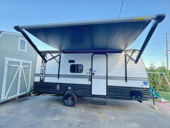 A bright blue sky over this RV rental travel trailer, featuring an extended awning with LED lights, parked next to a shed.. Keystone RV Hideout Sport 2024