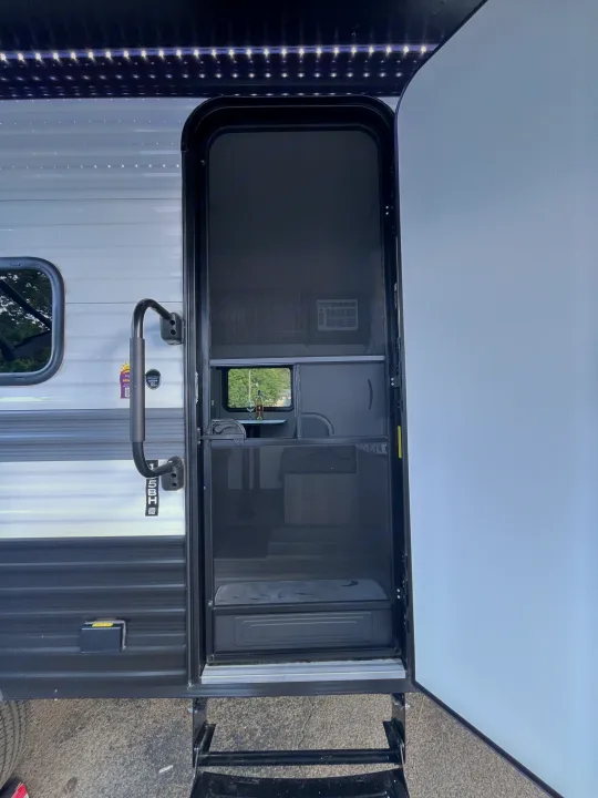 Open RV rental door with screen, entry steps, and interior showing a table, wine, and AC unit. Awning lights overhead.. Keystone RV Hideout Sport 2024