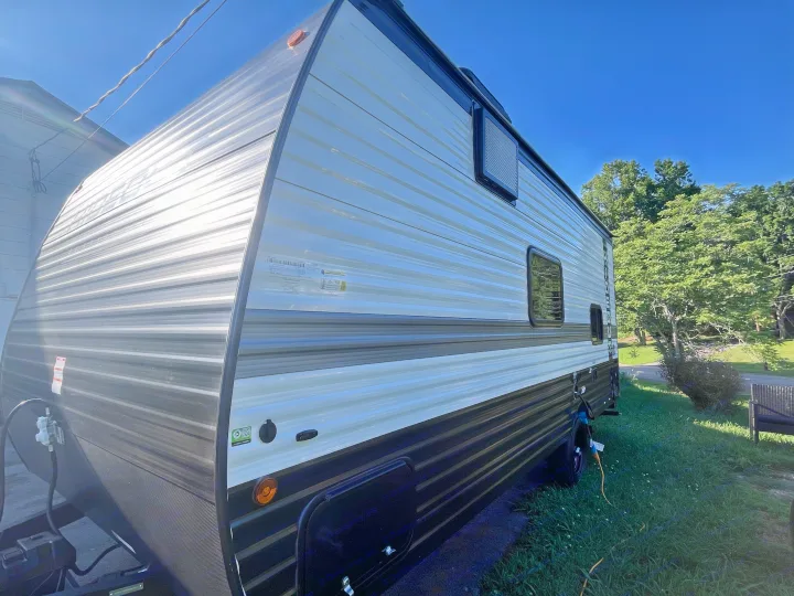 Modern RV rental parked on grass, showing its striped light & dark siding, windows, and power hookup on a sunny day.. Keystone RV Hideout Sport 2024