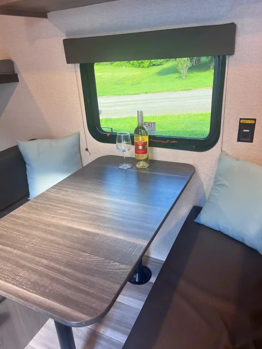 Inside an RV rental: a dark wood dinette table with wine and glass, light blue pillows, and a window view.. Keystone RV Hideout Sport 2024