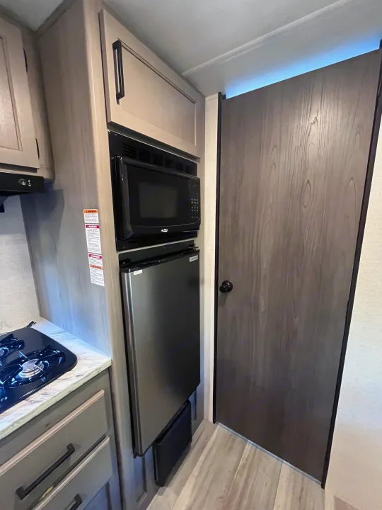 RV rental kitchen: a compact space with microwave, small fridge, and stovetop, plus wood-look cabinetry and door.. Keystone RV Hideout Sport 2024
