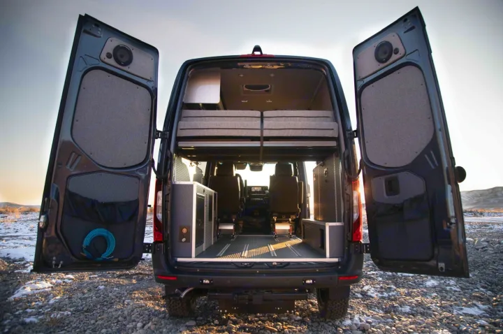 Rear view of a dark camper van with open doors, revealing a bed platform, modular storage, and a durable floor. Ready for RV rental adventures.. Mercedes Sprinter 2020