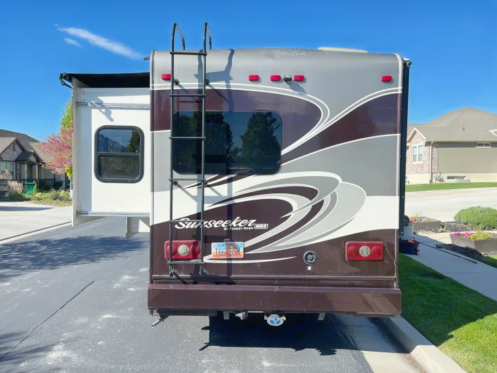 Rear view of a large, brown and gray RV with a ladder, extended slide-out, and camera. Parked on a residential driveway; a great rv rental option.. Forest River Sunseeker MBS 2016