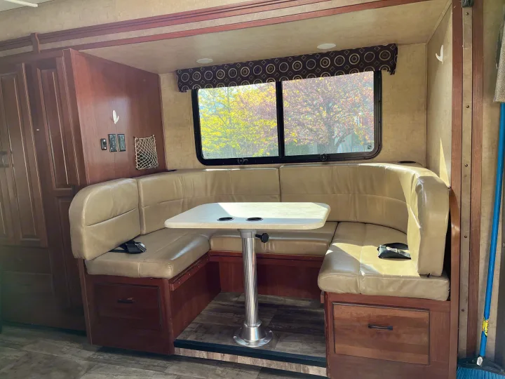 RV rental interior: a bright, U-shaped dinette with light cushions, center table, storage, and large windows revealing colorful trees.. Forest River Sunseeker MBS 2016
