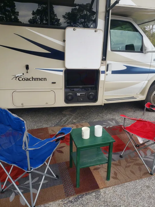 An RV rental provides an outdoor entertainment setup with a TV, sound system, green table, and two camping chairs on a rug.. Forest River Coachmen Leprechaun 2015