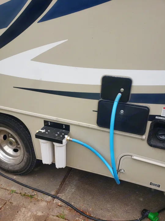 An RV rental is equipped with a dual-stage water filter and blue hose, connected to the vehicle for fresh water hookup and purification.. Forest River Coachmen Leprechaun 2015