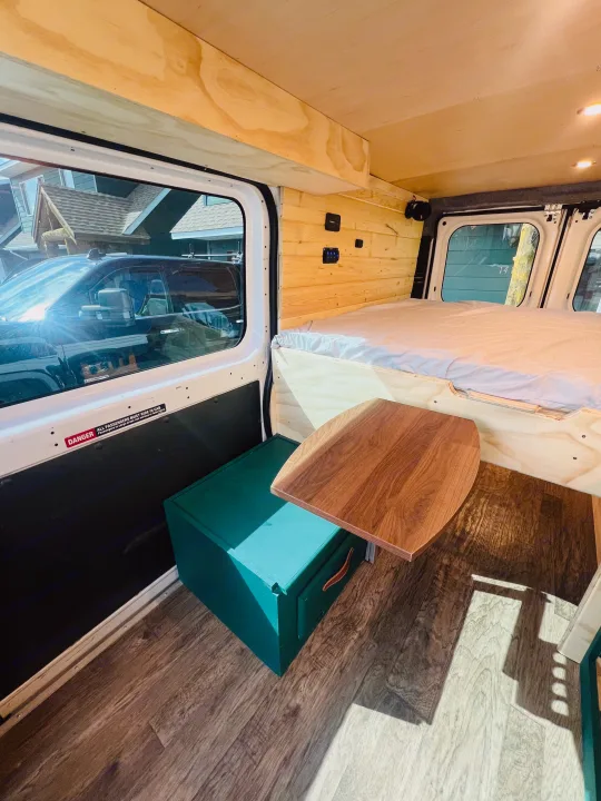 Experience an RV rental! This camper van interior features a bed, wood paneling, movable table, and green storage.. RAM Promaster 2017