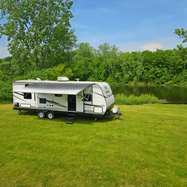 Enjoy nature with this **rv rental**! Travel trailer parked by a scenic river/lake surrounded by green trees & grass.. Jayco Jay Flight SLX8 2020