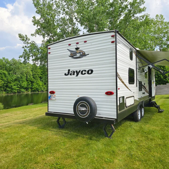 Jayco rv rental travel trailer parked on green grass next to a tranquil lake, surrounded by lush trees under a blue sky.. Jayco Jay Flight SLX8 2020