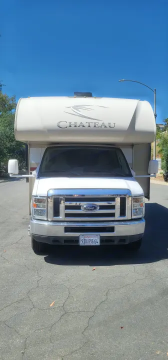 Front view of a white Ford Chateau Class C RV rental, parked on cracked asphalt under a bright blue sky.. Thor Chateau 2018