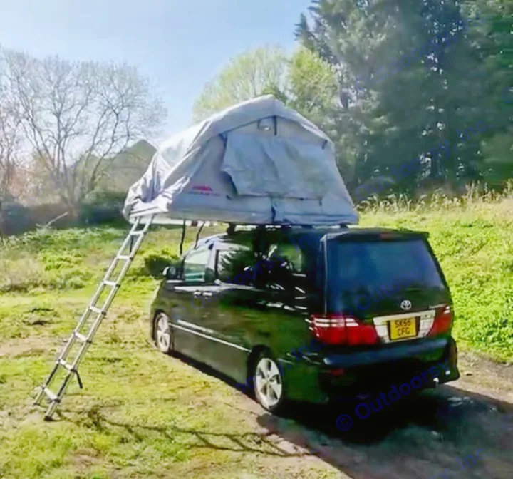 A black minivan with a rooftop tent and ladder is set up in a grassy field, ideal for an RV rental adventure.. Toyota Alphard 2024