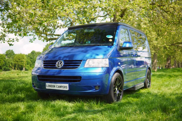 Blue London Campers VW camper van for RV rental, parked on sunny green grass amidst trees. Ideal for outdoor travel.. Volkswagen Other 2006