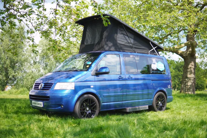 A blue VW pop-top campervan, an RV rental, sits on green grass with its roof extended. Trees surround it.. Volkswagen Other 2006