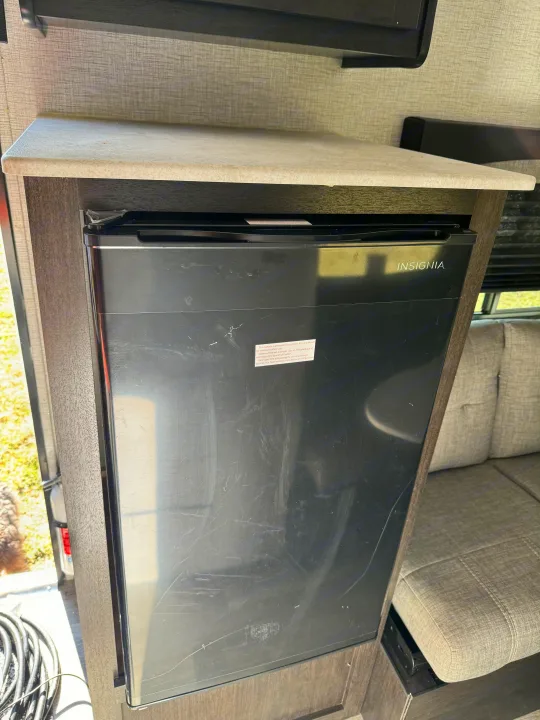 Compact Insignia refrigerator inside an RV rental, still with its protective film. A countertop is above, and a sofa is nearby.. Braxton Creek Bushwhacker Plus 2022
