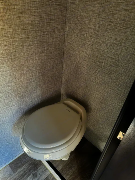 A white Dometic toilet in the compact bathroom of an RV rental, featuring textured gray walls and a grey wood-look floor.. Braxton Creek Bushwhacker Plus 2022