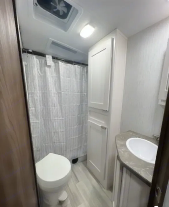 Clean RV rental bathroom with toilet, striped shower curtain, sink vanity, and tall storage cabinet, featuring modern light finishes.. Cruiser RV MPG 2023