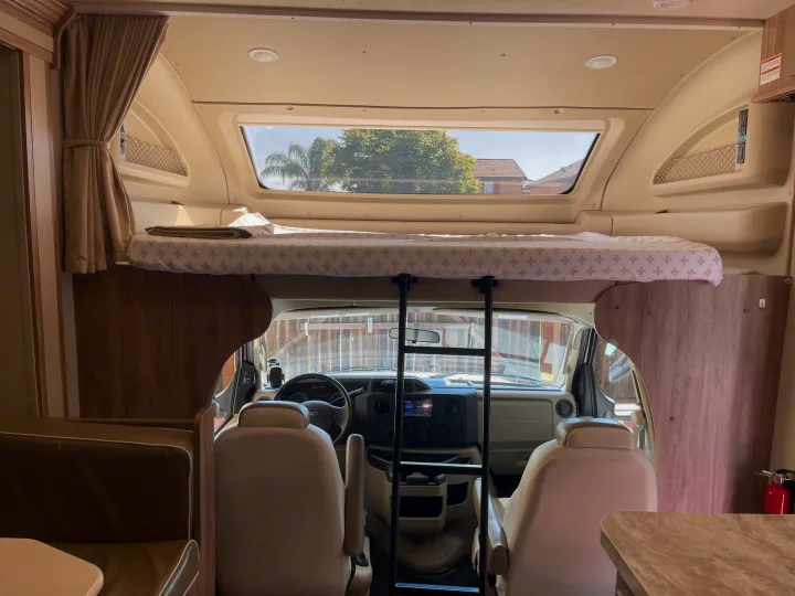 Bright RV rental interior shows a neatly made overhead bunk bed, ladder, and front cockpit area with seating.. Entegra Coach Odyssey 2019