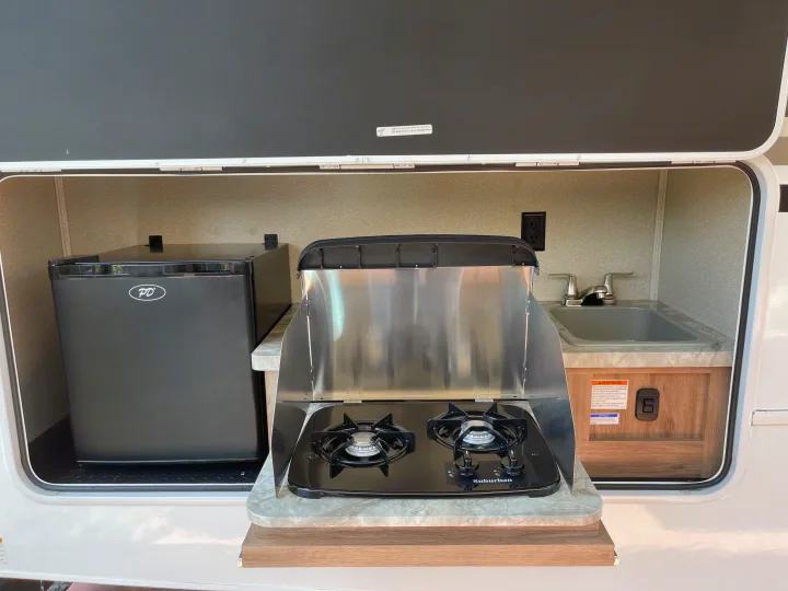 Convenient outdoor kitchen in an **rv rental**, featuring a compact PD fridge, Suburban 2-burner stovetop, and a small sink.. Entegra Coach Odyssey 2019