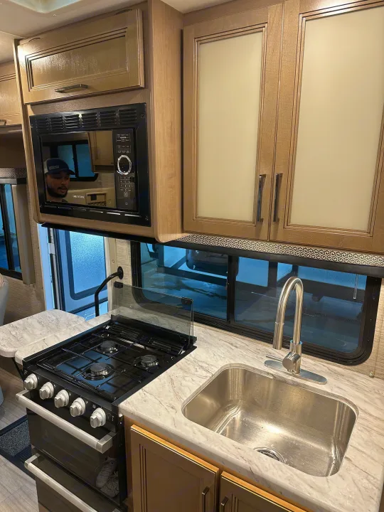 Spacious RV rental kitchen featuring a microwave, stove, sink with a tall faucet, and light wood cabinetry.. Thor Coleman 2021