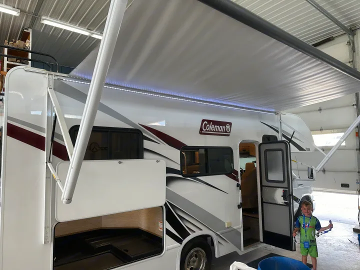 A white RV with its awning & LED lights extended, and an open storage bay. A young boy stands by the entry, ready for an rv rental adventure.. Thor Coleman 2021