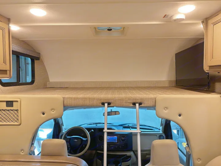 Inside an **RV rental**: the comfy cab-over bunk with a TV, overlooking the cockpit with steering wheel and controls.. Thor Coleman 2021