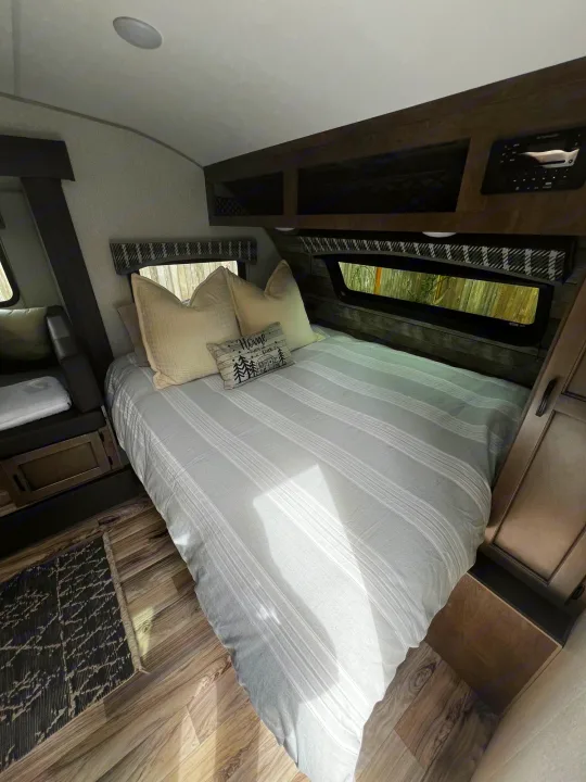 Cozy RV rental bedroom featuring a striped bed, overhead storage, and a bench area, ideal for mobile living.. Sonic Lite 2022