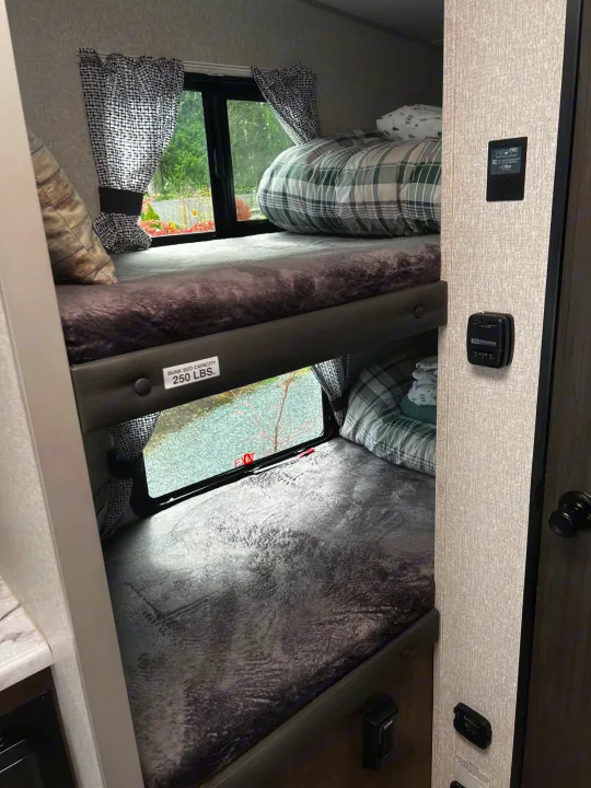 Inside an RV rental: view of stacked bunk beds with plaid and solid blankets, windows, and wall-mounted controls for comfort.. Sonic Lite 2022