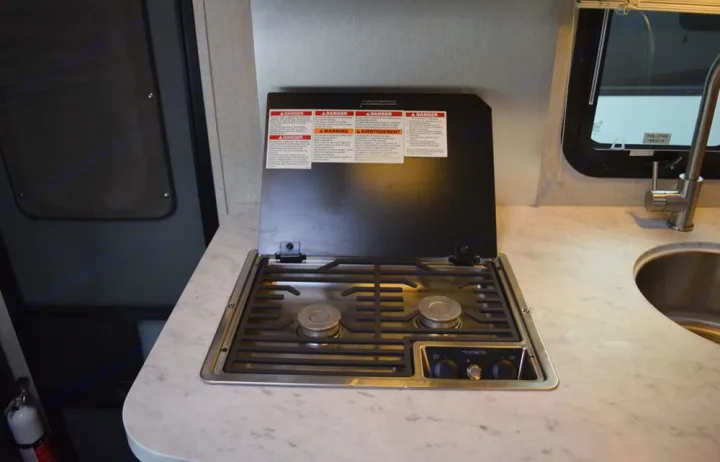 Compact RV rental kitchen features a Dometic two-burner stovetop with safety warnings on its cover, alongside a stainless steel sink.. Adventurer Adventurer 2020