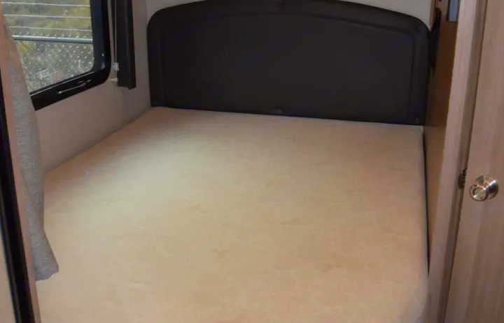 RV rental bed features a light mattress, dark headboard, window, and nearby wood door, ready for a comfortable stay.. Adventurer Adventurer 2020