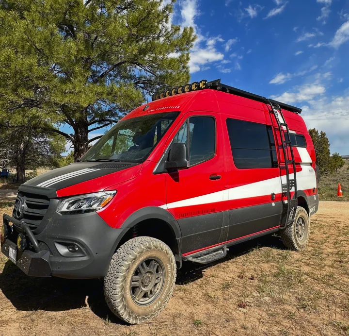 A vibrant red Storyteller Overland camper van, equipped for off-road adventures, parked among pine trees. Ideal for an RV rental.. Storyteller Overland Beast Mode AWD 2024