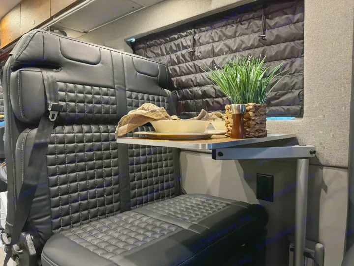Cozy RV rental interior features quilted black seating, a folding table with a plant and meal setup, and insulated window cover.. Storyteller Overland Beast Mode AWD 2024