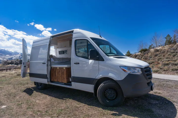 This white RV rental camper van is parked outdoors with its doors open, revealing its custom interior conversion.. Mercedes-Benz Sprinter-Loaded 2022