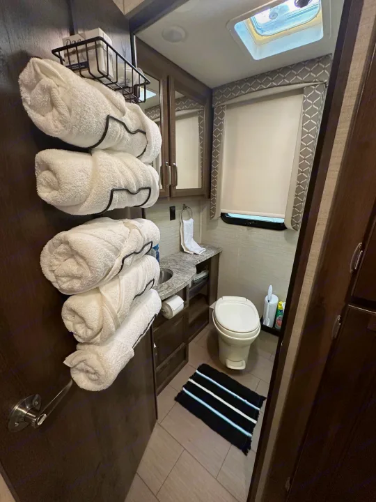 Inside this RV rental, a compact bathroom features a toilet, sink, and neatly stacked white towels for guests.. Jayco Greyhawk 2019