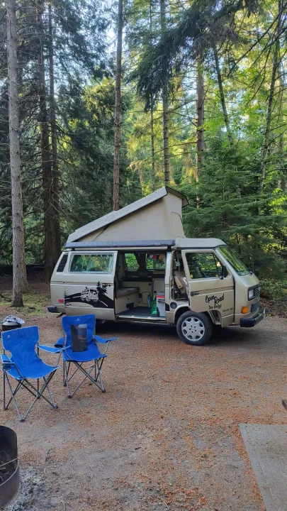 Relaxing at a forest campsite with a tan camper van from Open Road RV rental. Chairs and fire pit ready.. Volkswagen Westfalia 1989