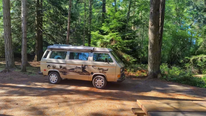 A beige VW camper van, an RV rental from Open Road Van Rentals, is parked on a dirt path in a sunlit forest campsite.. Volkswagen Westfalia 1989