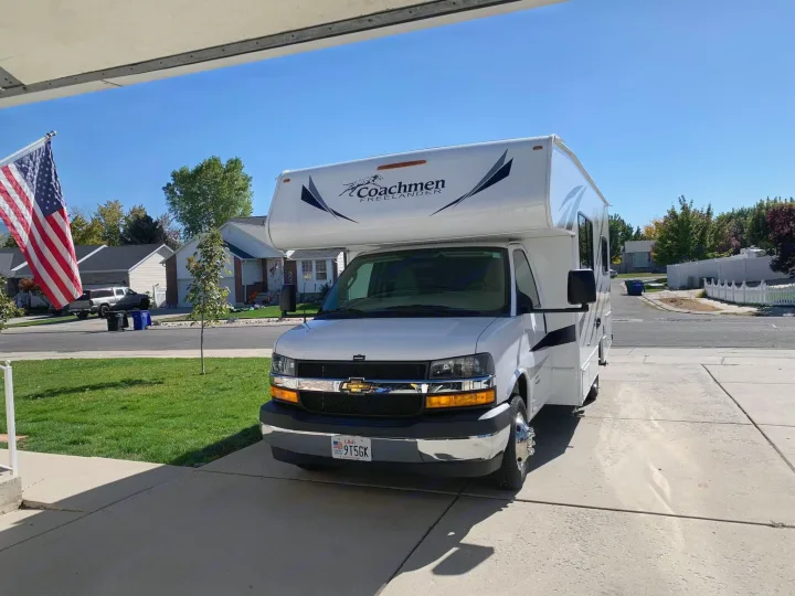 White Coachmen Freelander Class C motorhome (RV rental) parked in a sunny driveway. An American flag is visible on the left.. Coachmen Freelander 2020
