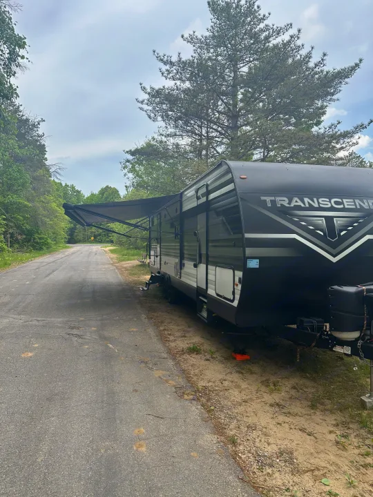 Black Transcend RV rental parked on a paved road in a wooded setting with its awning extended, ready for camping.. Grand Design Transcend Xplor 2022