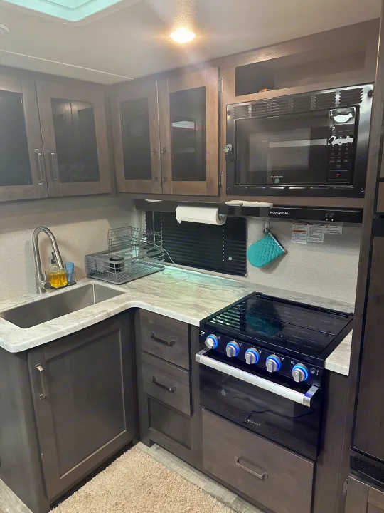 This RV rental kitchen offers a compact, modern design with a sink, microwave, stove, and sleek dark wood cabinets.. Grand Design Transcend Xplor 2022