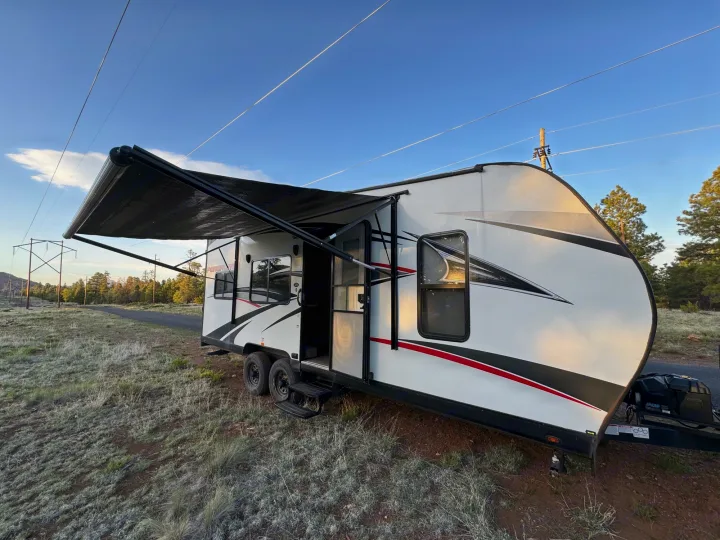 White RV rental travel trailer with awning extended, parked off-road in frosted grass, under power lines. Perfect for camping.. Pacific Coachworks Blaze'N 2017