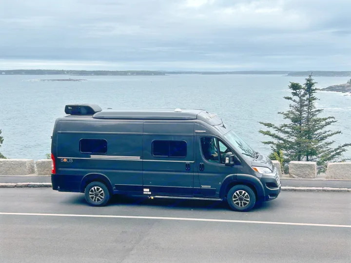 This dark Winnebago Solis camper van rv rental is parked oceanside on a cloudy day, offering stunning coastal views for road trip fun.. Winnebago Solis 2023