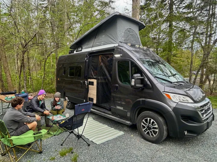 Three people play a board game outside their pop-top RV rental camper van at a wooded campsite.. Winnebago Solis 2023