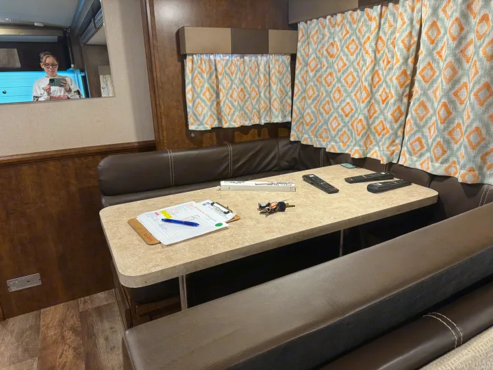 RV rental dinette featuring brown booth seating, patterned curtains, and a table with various items. Person visible in mirror.. Forest River FR3 2016