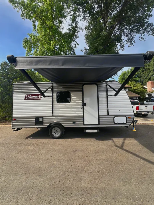 This Coleman RV rental, a white and gray travel trailer with its black awning extended, is parked outdoors on asphalt.. Coleman Lantern 17B 2024