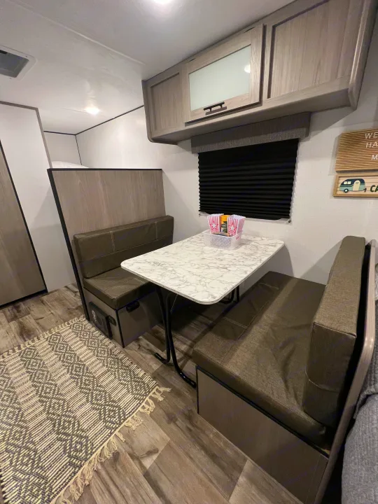 Step inside this modern RV rental! Features a dinette with upholstered benches, a marble-look table, and overhead cabinets. Bunk bed visible.. Coleman Lantern 17B 2024